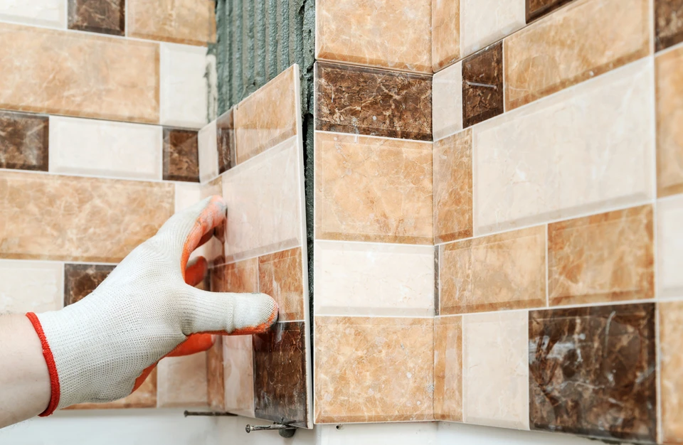 Une main gantée installe des carreaux de céramique sur un mur d'angle, créant un motif décoratif avec des tons beiges, marrons et blancs