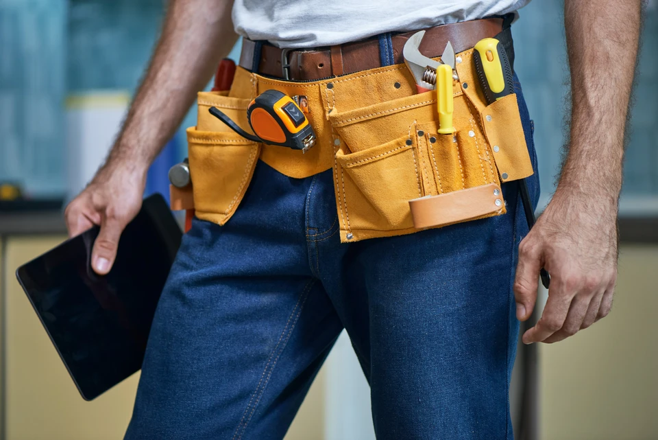 Artisan en jeans et ceinture porte-outils en cuir travaillant à l'intérieur, tenant une tablette numérique noire
