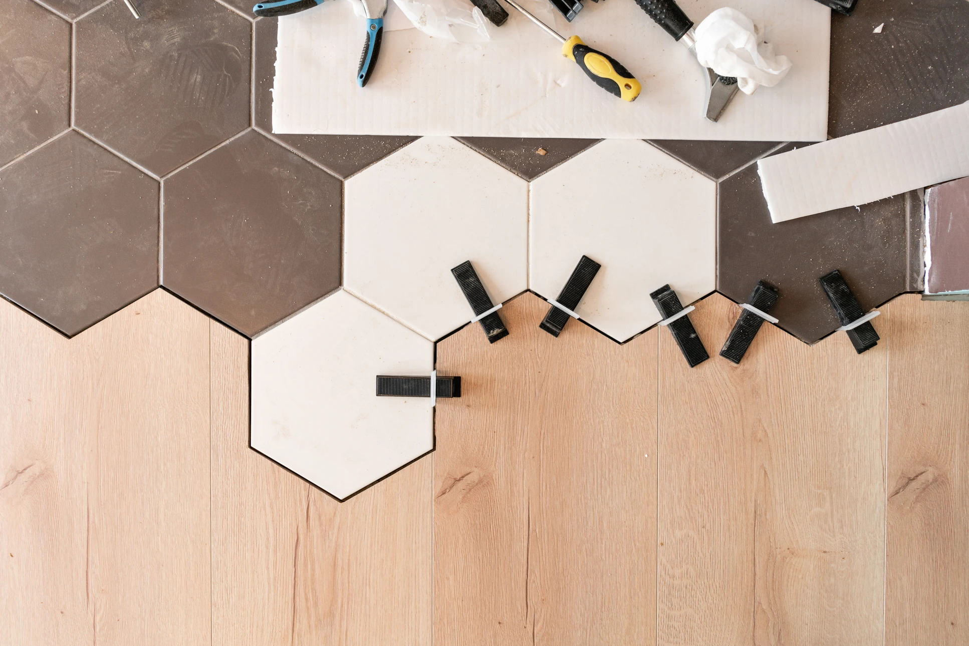 Plan rapproché d'un artisan qualifié en train de poser du carrelage au mur dans une salle de bains moderne. L'artisan porte des équipements professionnels (gants, lunettes de protection) et utilise un niveau et une spatule pour assurer l'alignement parfait des carreaux. Les carreaux sont partiellement posés, montrant la progression du travail avec précision. L'arrière-plan révèle une salle de bains en cours de rénovation avec des murs préparés. La lumière naturelle illuminate le travail soigné et méticuleux, mettant en évidence la qualité de l'exécution et l'expertise de l'artisan.